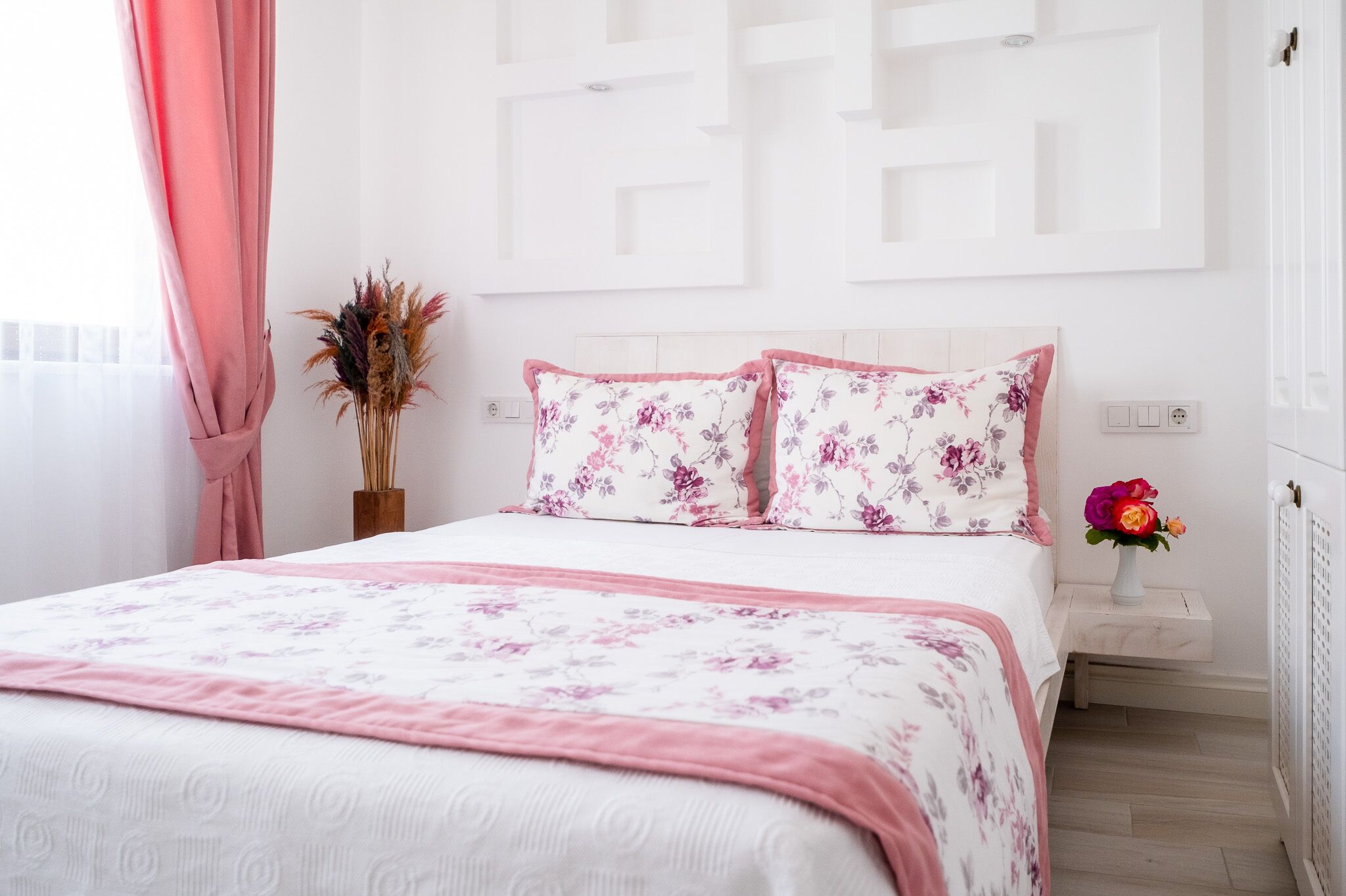 A neatly arranged bedroom with a bed, floral bedding, a vase with dried flowers, and a small table with a vase of fresh flowers.
