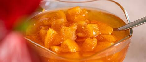 Uma tigela de vidro transparente cheia de uma sobremesa com fruta semelhante a pêssego, colocada sobre uma mesa com um pão em segundo plano.