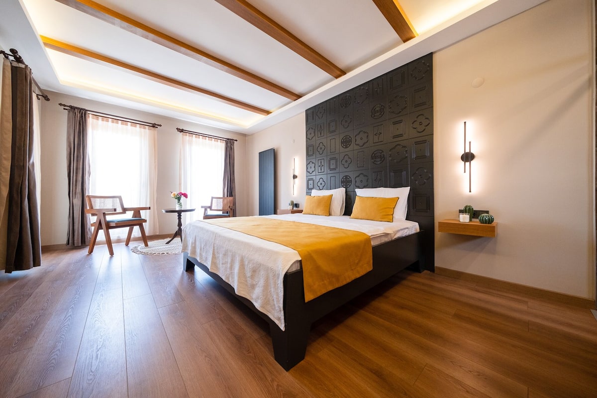 A modern hotel room with a wooden floor, a bed with a white and yellow bedspread, a desk with a chair, and a wall-mounted shelf with decorative items.