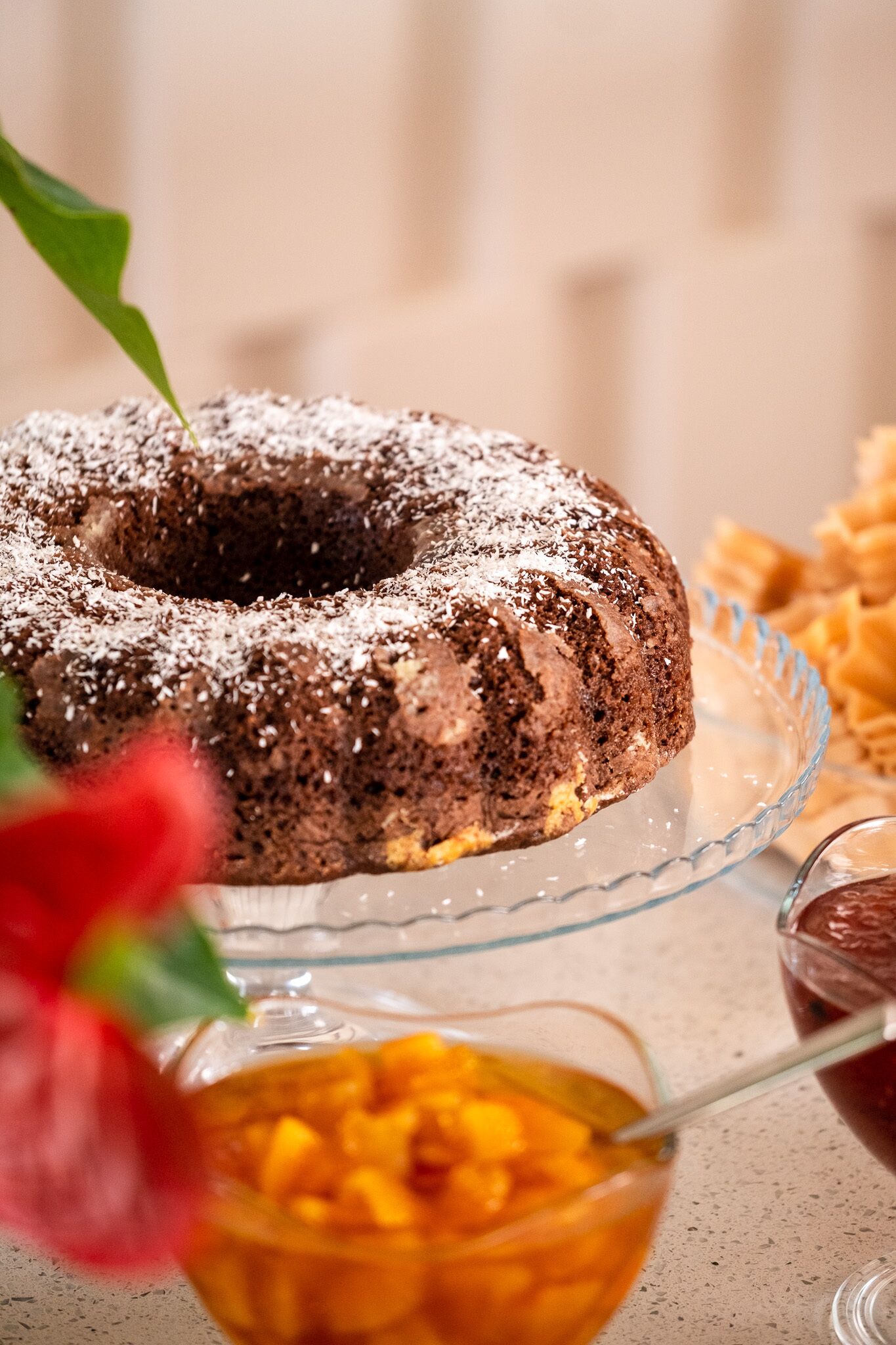 Un gâteau au chocolat en forme de couronne, présenté sur une assiette en verre, accompagné d’une cuillère et d’un bol de fruits.