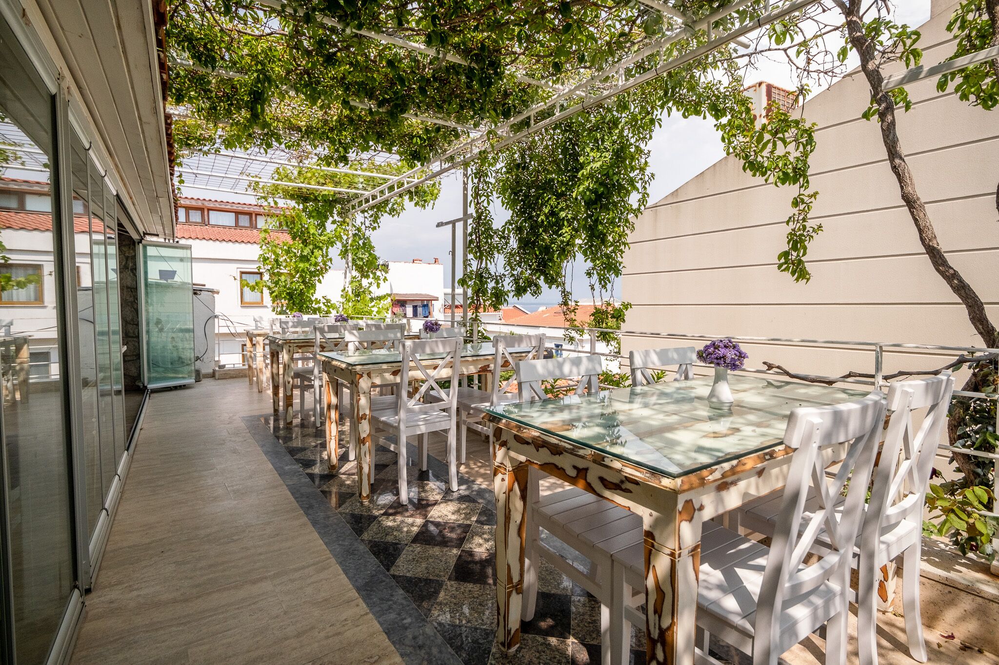 Un patio al aire libre con mesas de vidrio y sillas blancas, una pérgola arriba y vista de edificios al fondo.