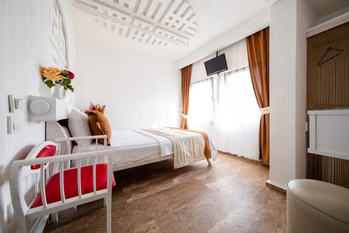 A small, well-lit room with a single bed, a white chair, a small table, and a TV mounted on the wall.
