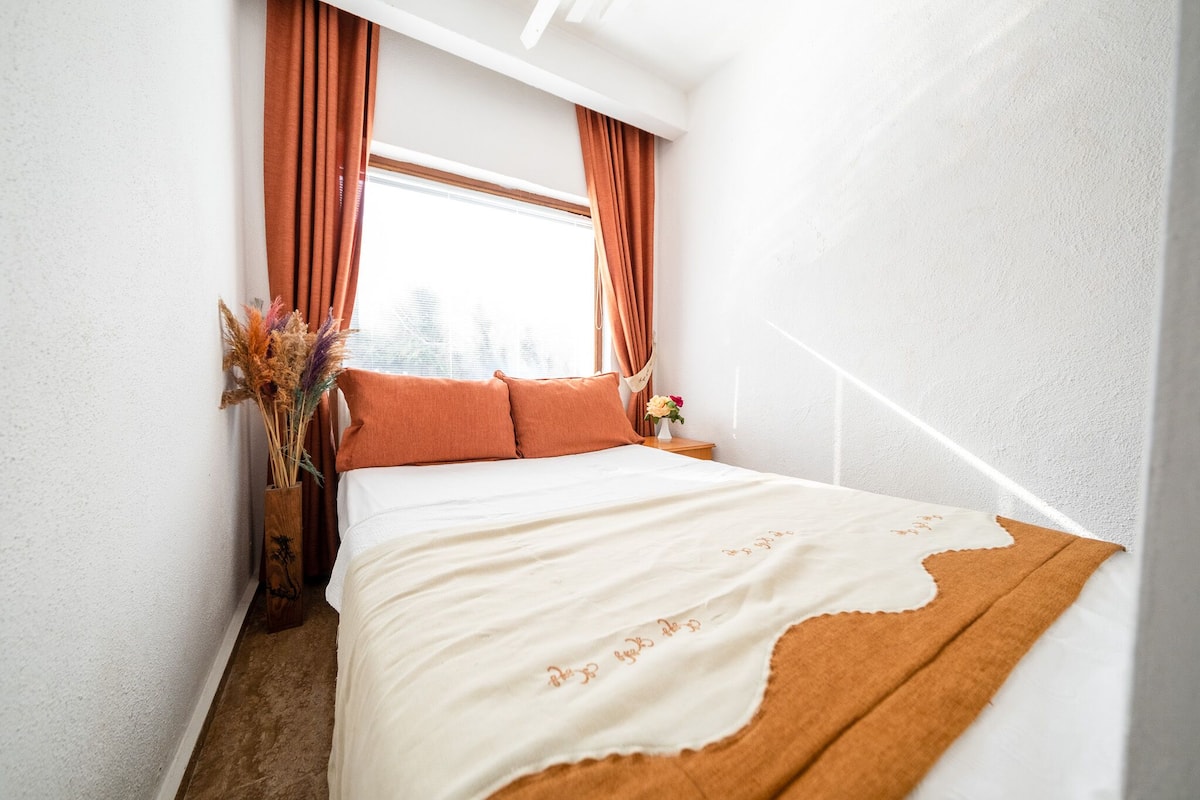 A bedroom with a bed, orange curtains, a window, and a vase with dried plants.