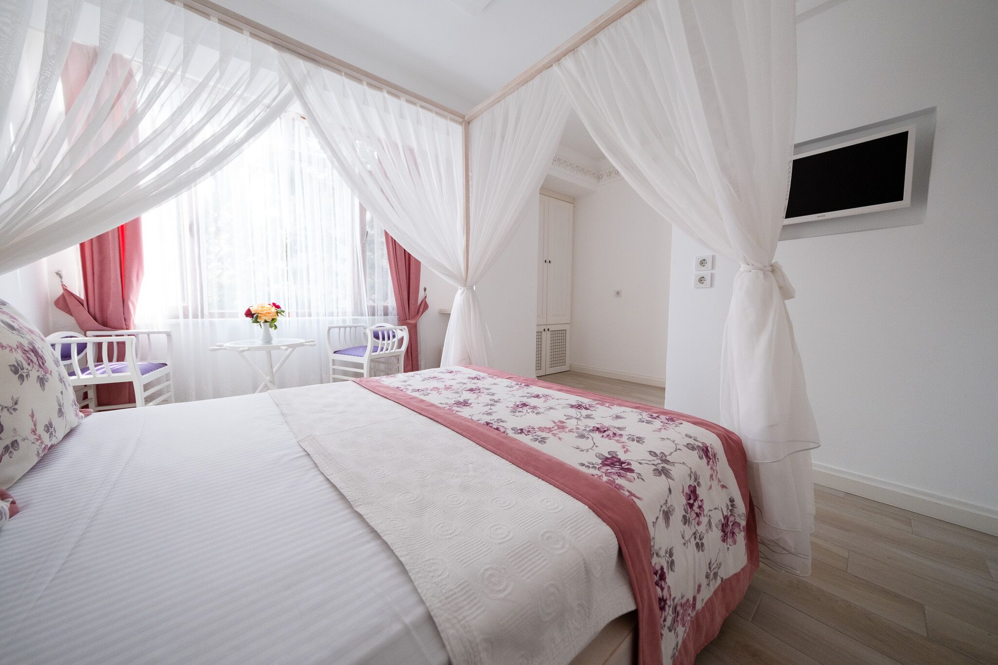 A bedroom with a bed, a small table, and a chair. There is a window with curtains and a flower vase.