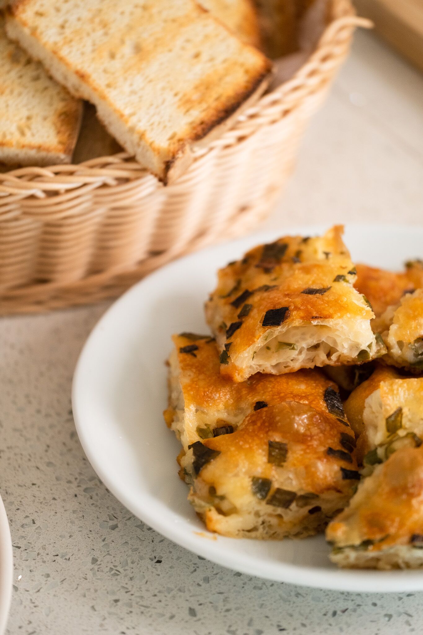 A plate of golden-brown baked goods with visible herbs, accompanied by a basket of plain bread slices in the background.