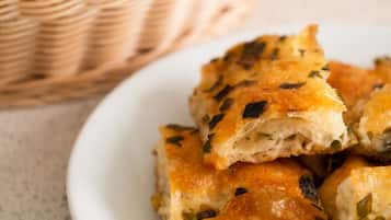 A plate of golden-brown baked goods with visible herbs, accompanied by a basket of plain bread slices in the background.
