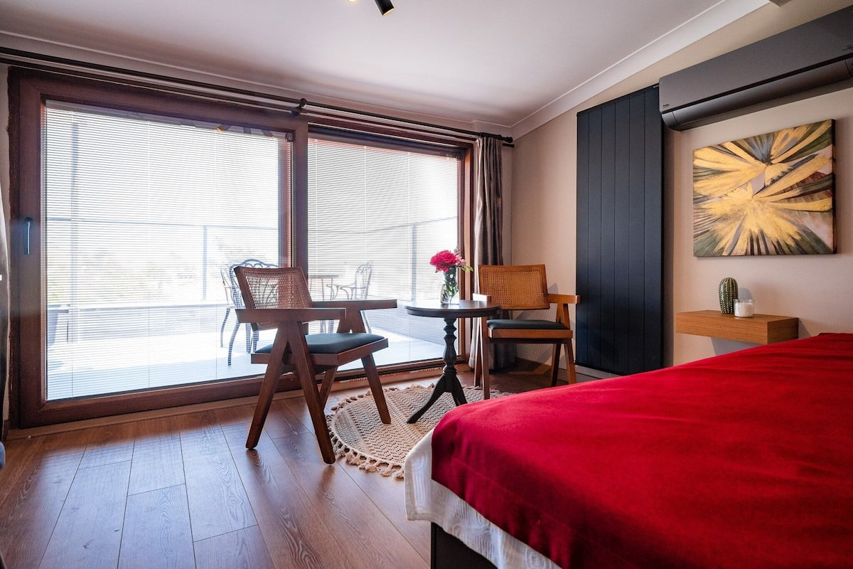A hotel room with a large window, a bed with a red blanket, a wooden chair, a small table with a vase of flowers, and a wall-mounted air conditioner.