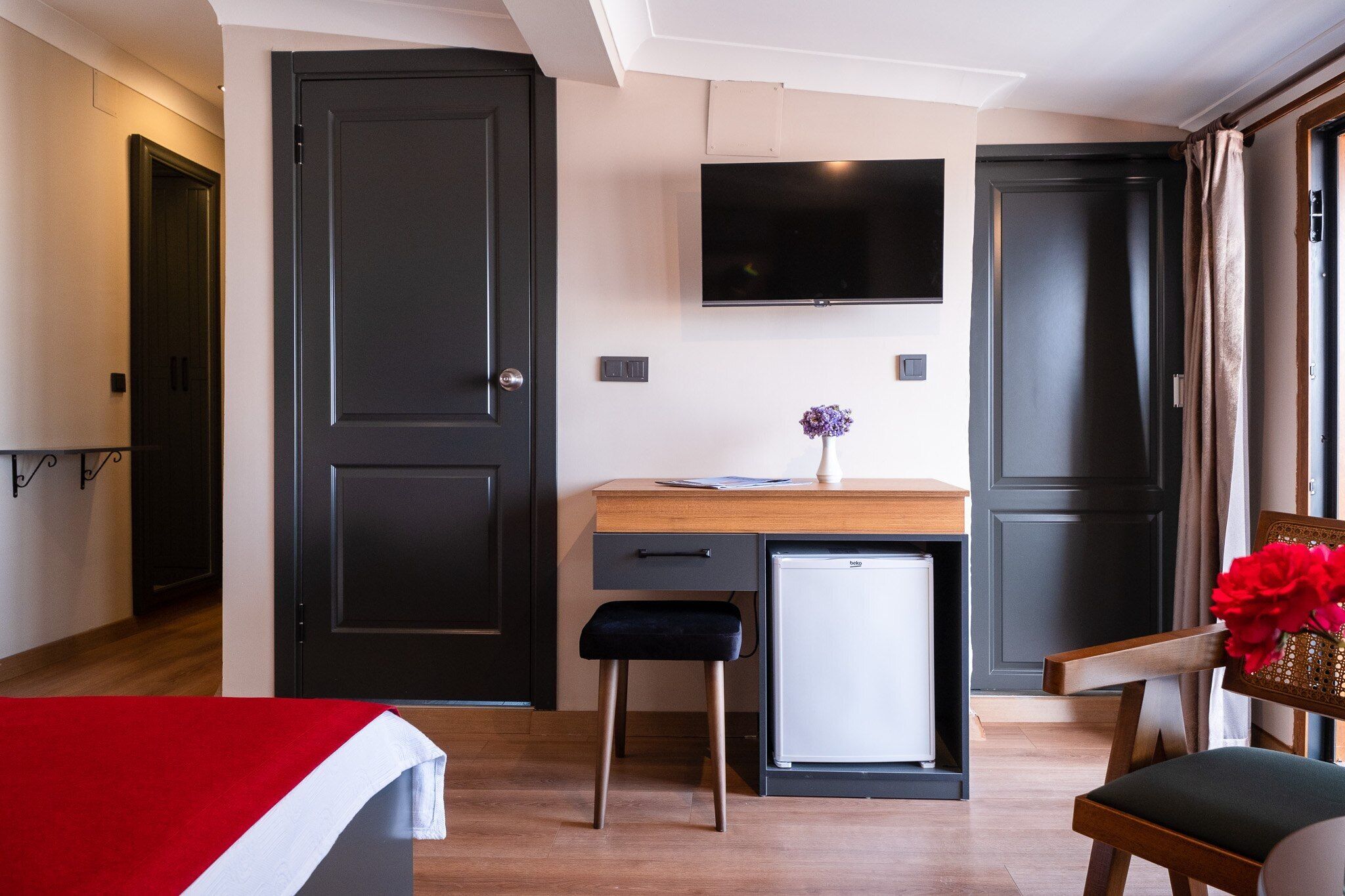 Habitación funcional con cama, escritorio con computadora, televisor montado en la pared y refrigerador pequeño.