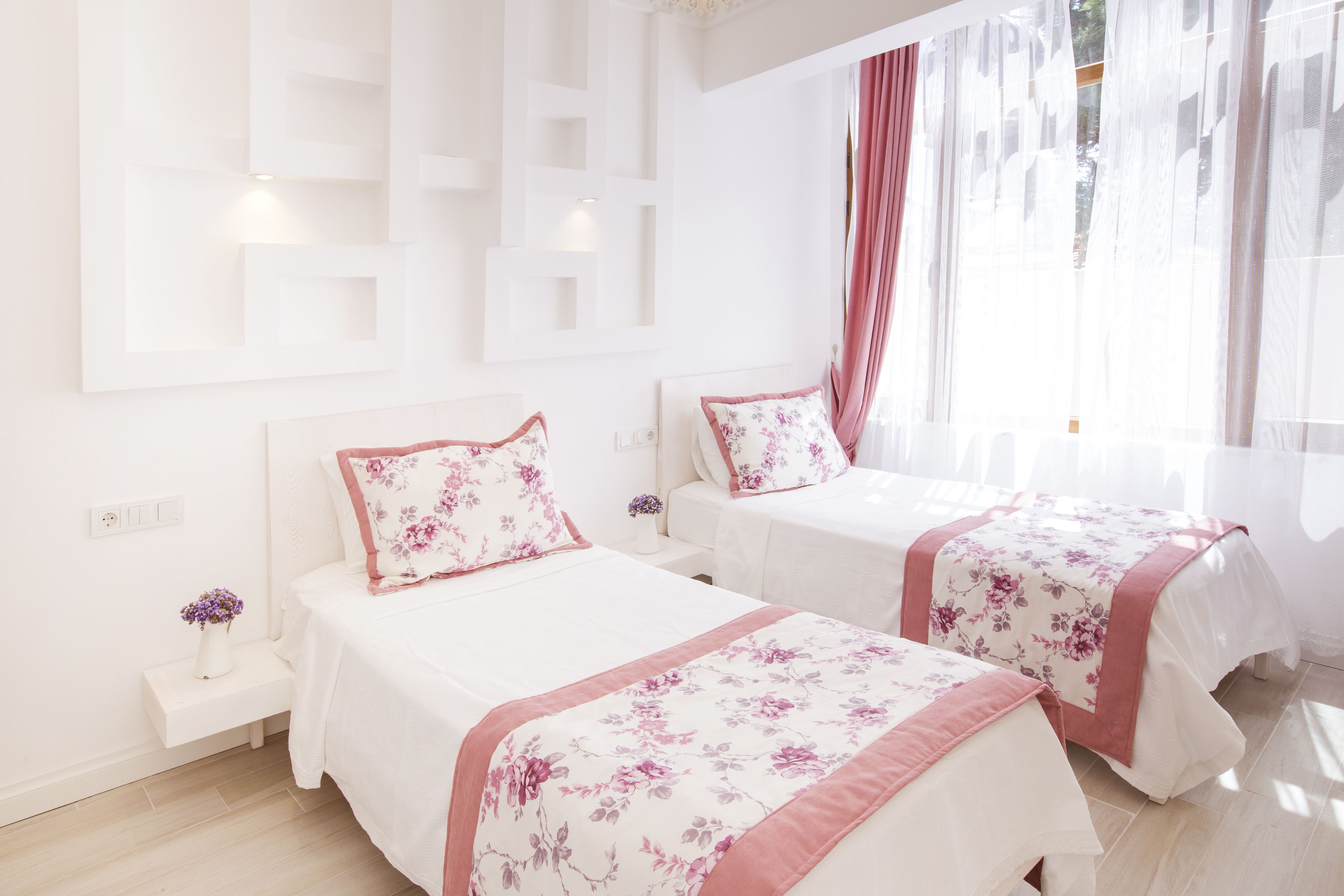 A hotel room with two beds, each with floral bedding and pink accents, a bedside table with a vase, and a large window with sheer curtains.