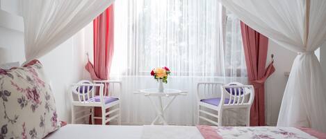 A bedroom with a bed, two chairs, a small table, and a window with sheer curtains.