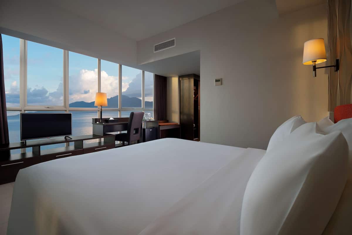 A hotel room with a large bed, a desk, a chair, a television, and a view of the sea through large windows.