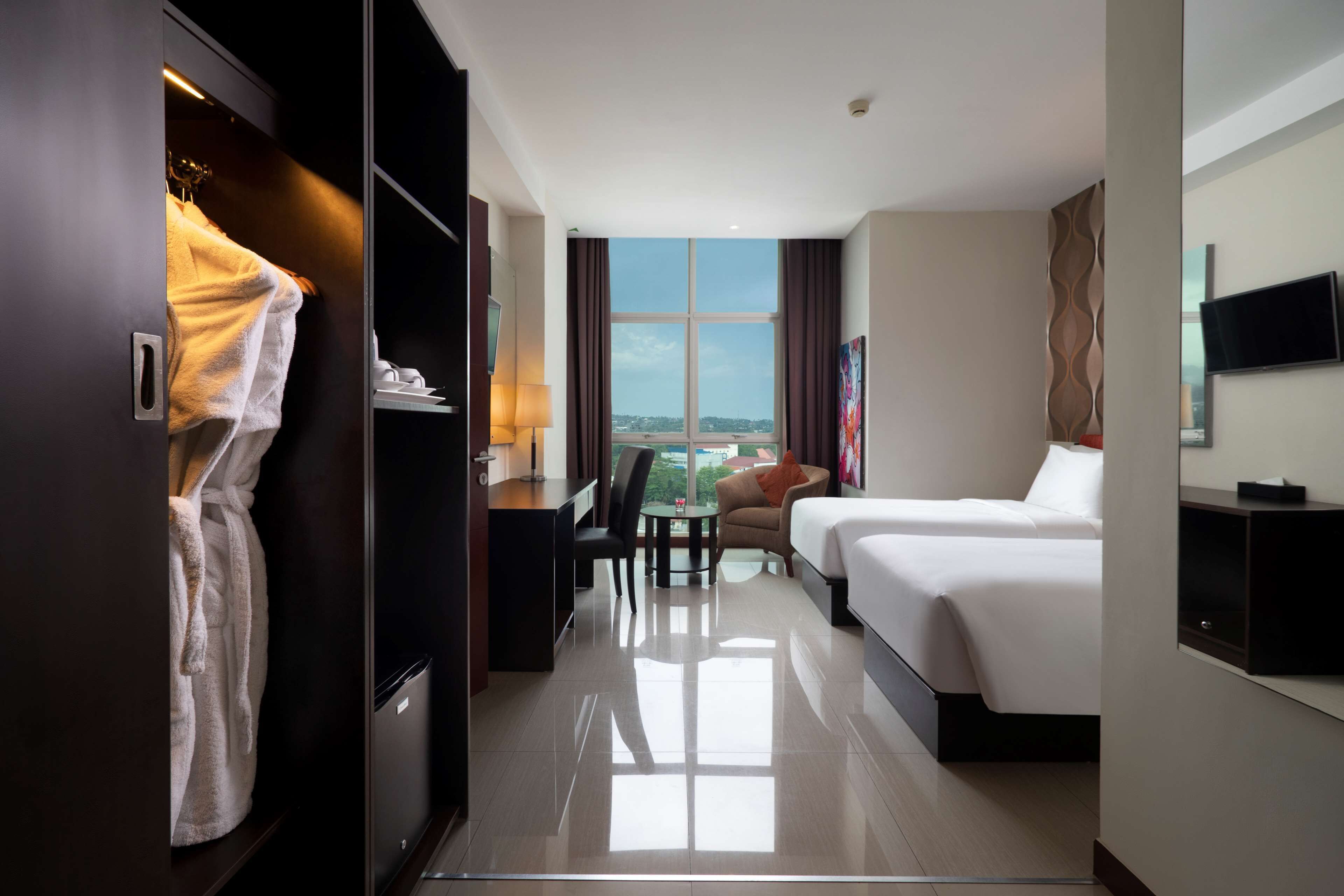 Habitación de hotel con cama, escritorio y silla, una mesita, televisor y ventana grande con vistas a la ciudad.