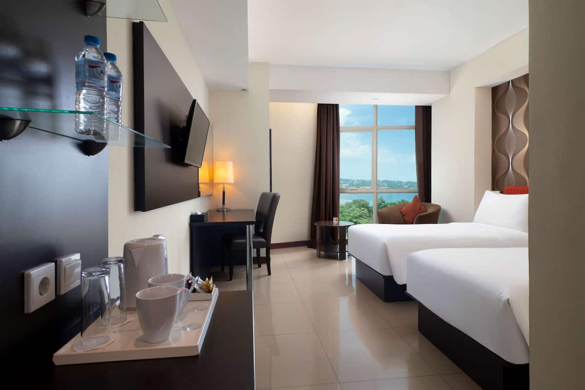 A hotel room with a bed, a desk with a chair, a TV mounted on the wall, a window with a view, and a tray with cups and a water bottle.