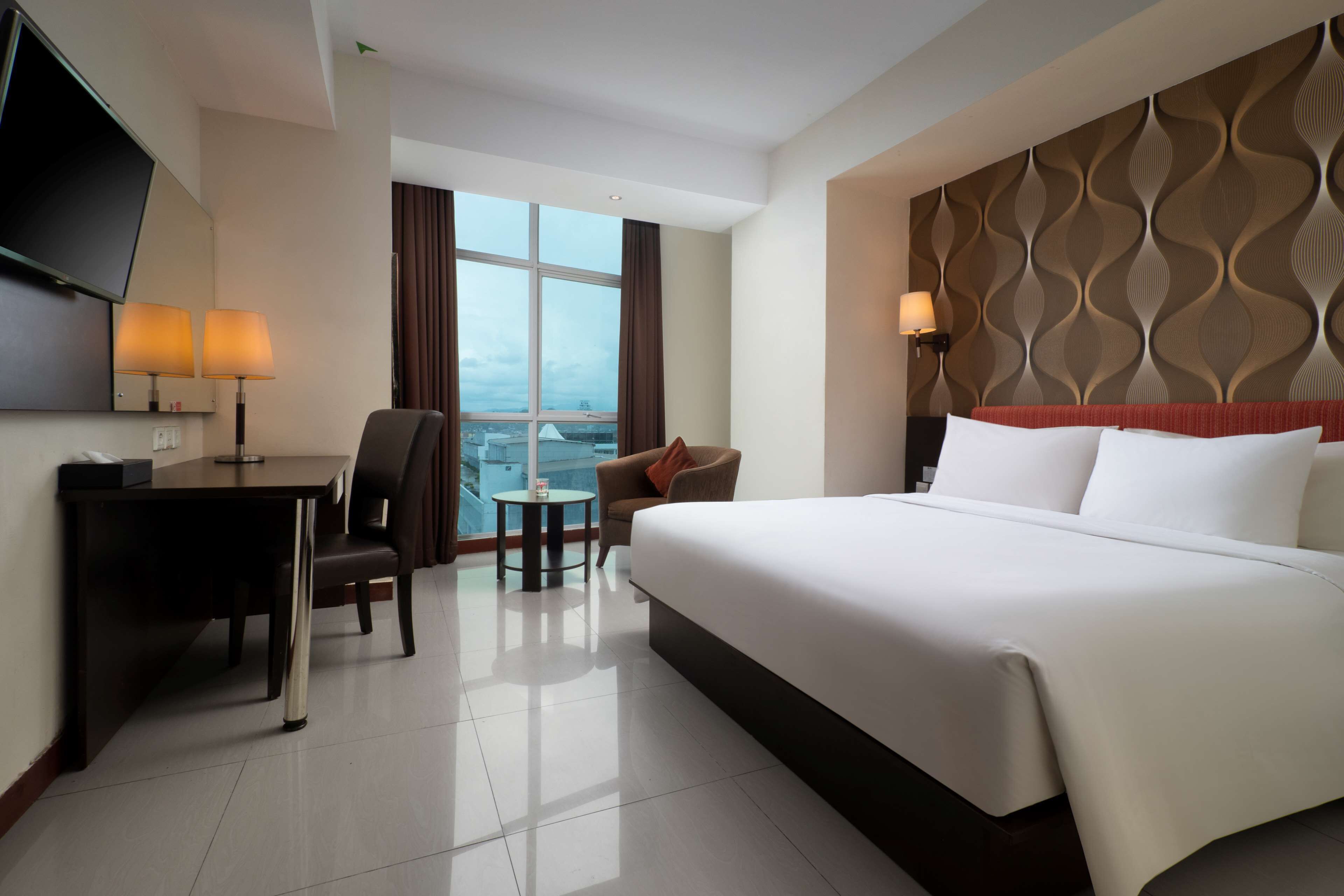A hotel room with a large bed, a desk with two lamps, a chair, a small table, and a TV mounted on the wall.