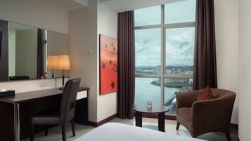 A hotel room with a desk, chair, bed, and a view of a body of water through large windows.