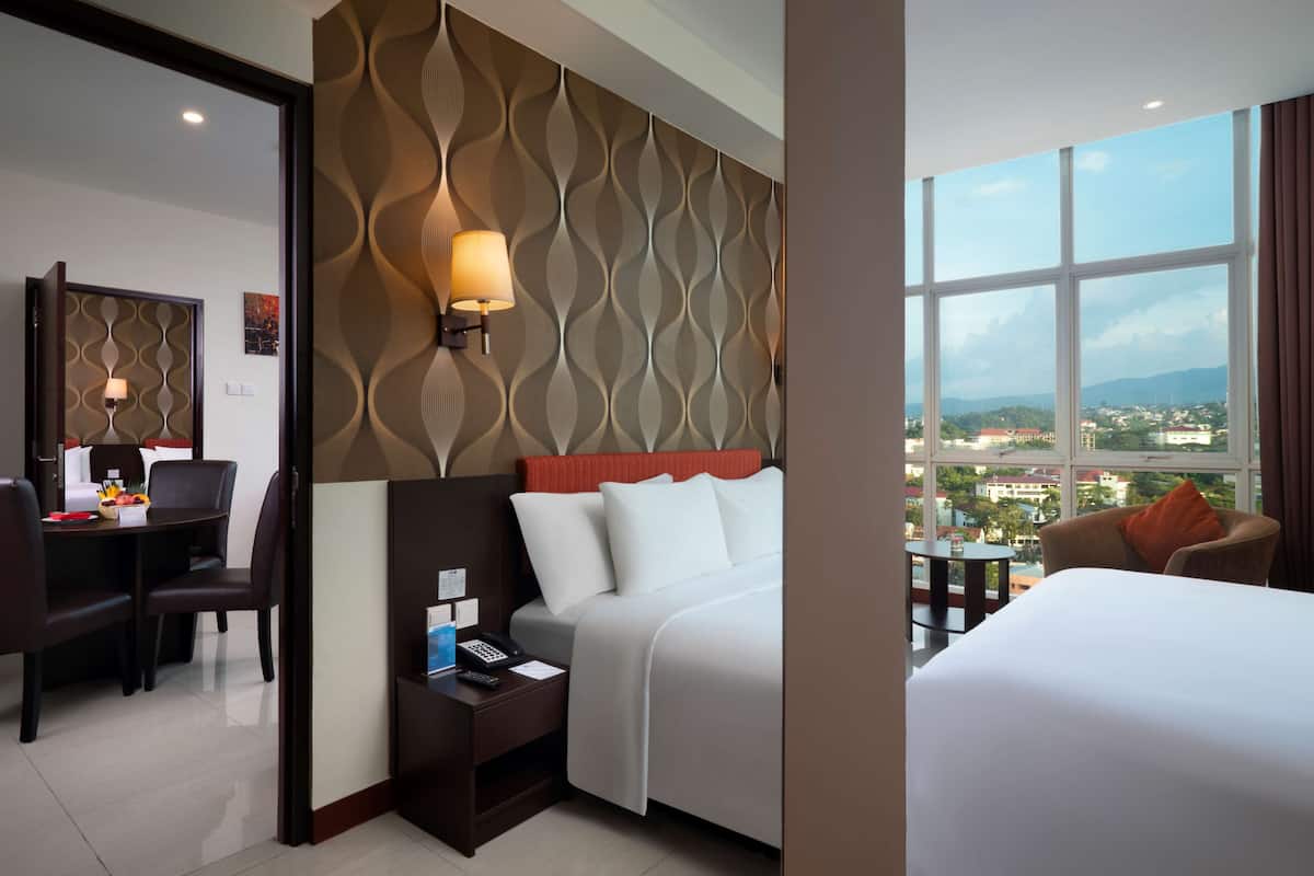 A hotel room with a bed, a desk, a chair, a lamp, and a view of the city through large windows.