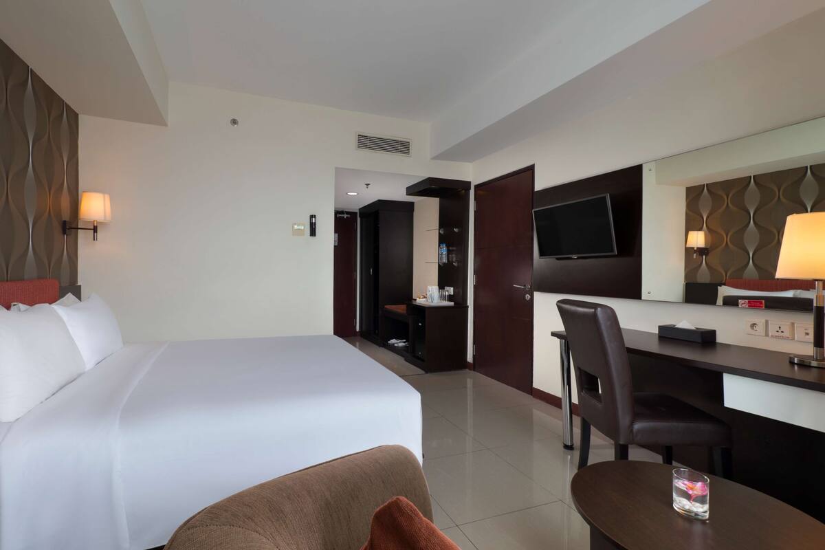 A hotel room with a large bed, a desk with a chair, a TV mounted on the wall, and a mirror above the desk.