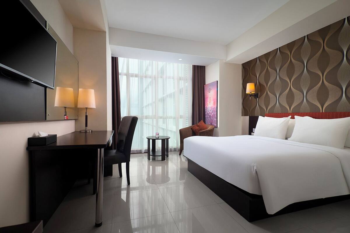 A hotel room with a bed, desk, chair, TV, and a view of the city.
