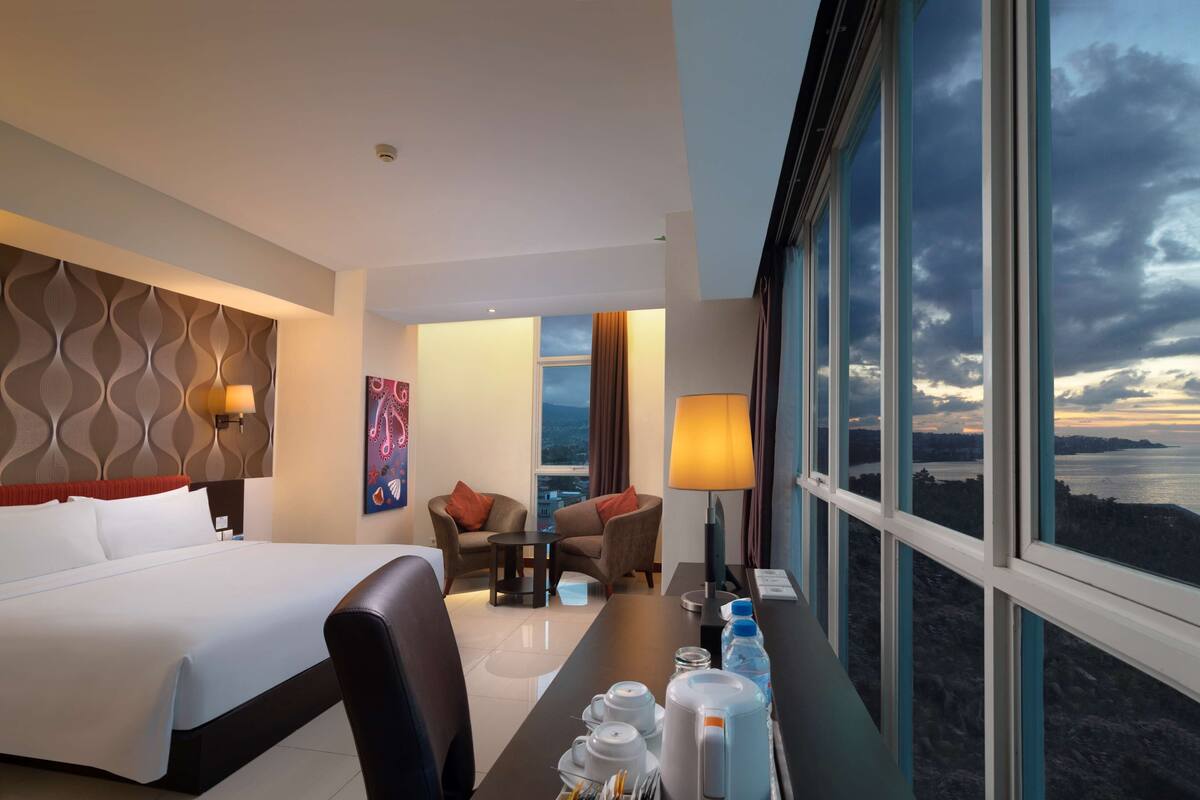 A hotel room with a bed, two armchairs, a small table, a desk with amenities, and large windows offering a view.