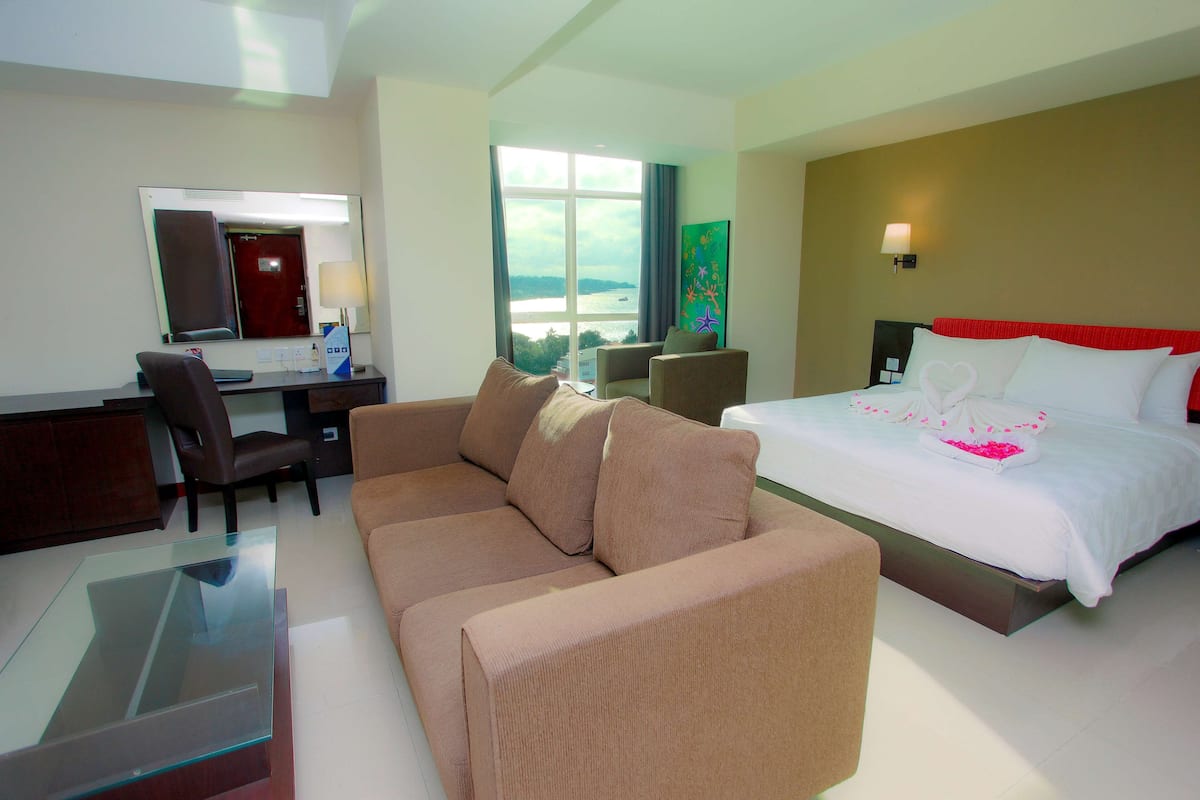A hotel room with a bed, a sofa, a desk, and a large window offering a view of the sea.