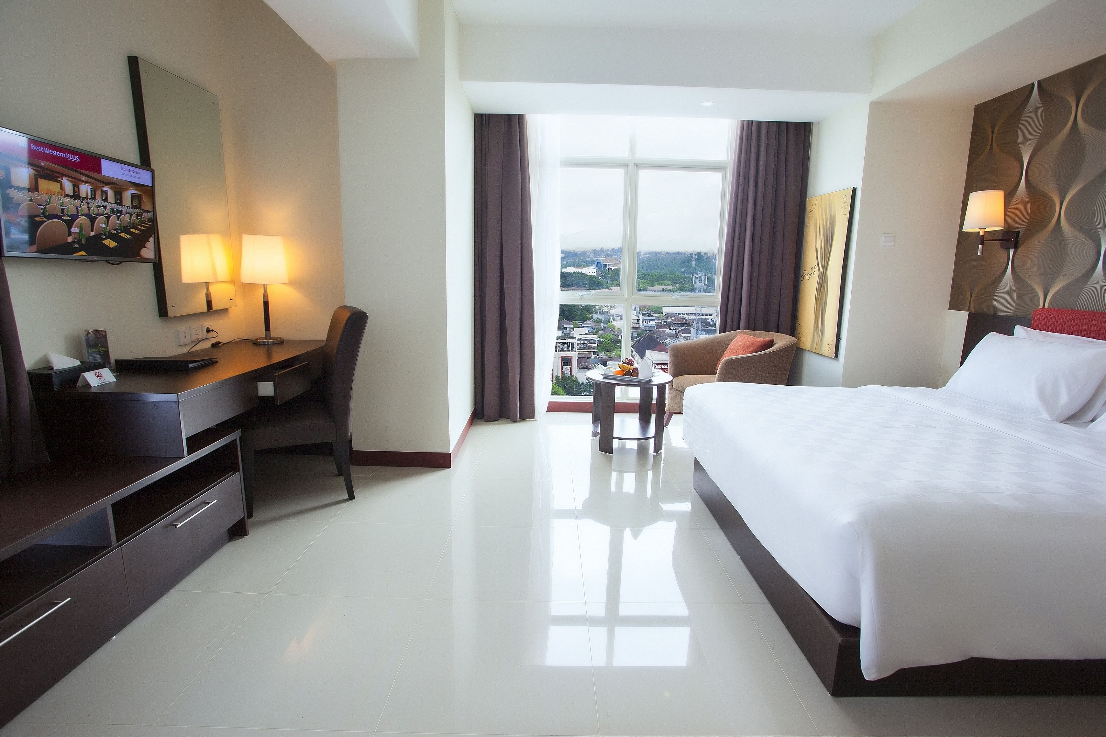 A hotel room with a bed, desk, chair, and a view of the city through large windows.