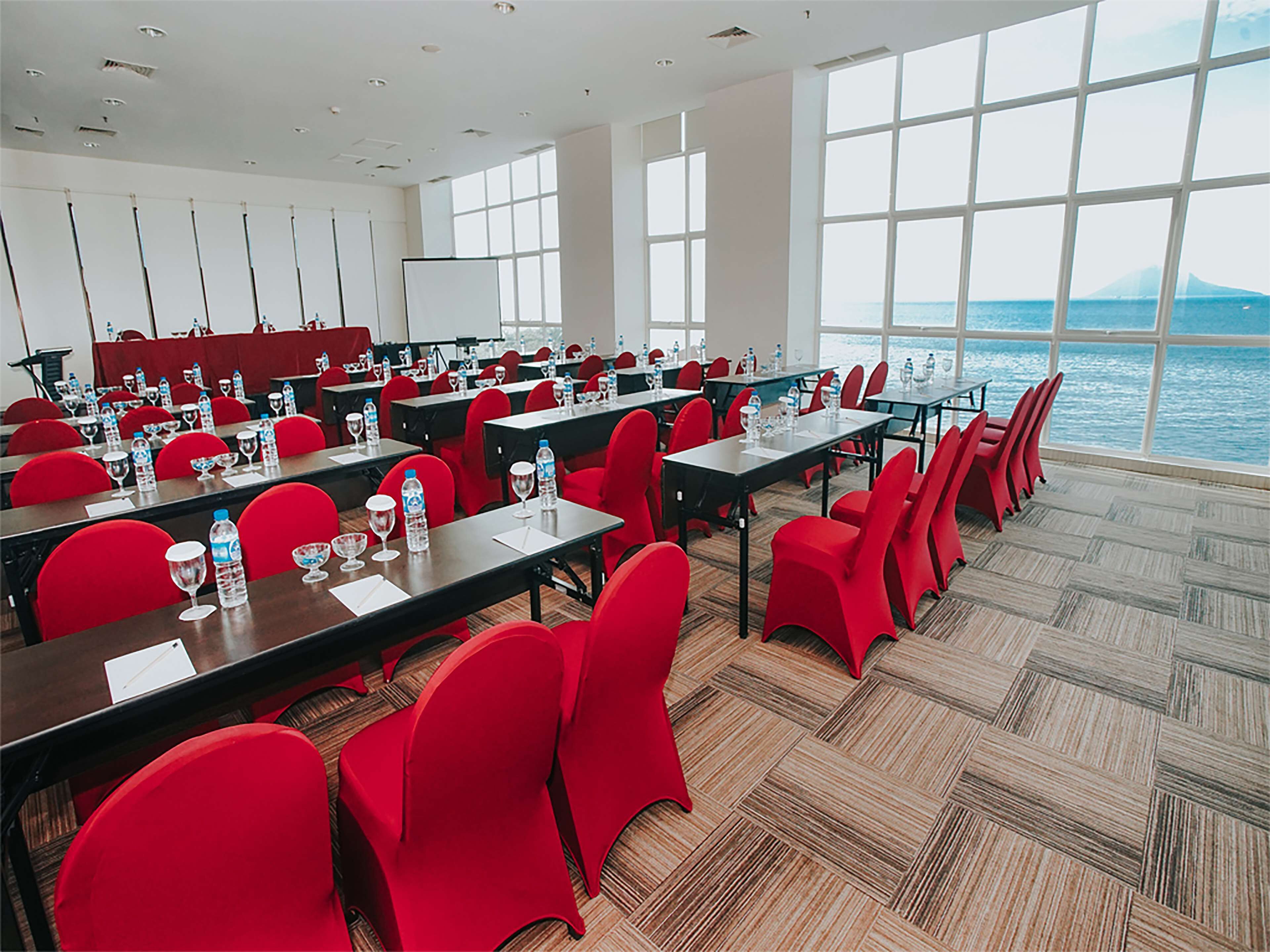 A conference room with red chairs and tables, large windows, and a view of the sea.