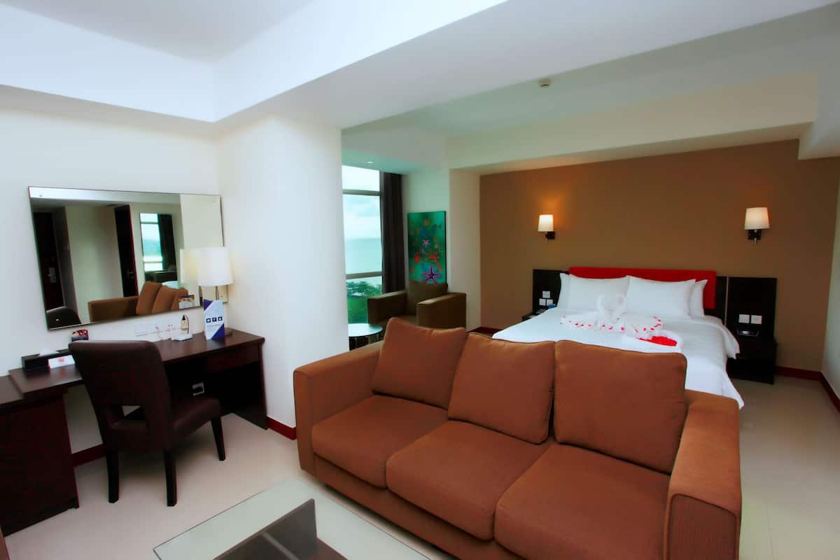 A hotel room with a bed, a brown sofa, a desk with a chair, a mirror, and a painting on the wall.