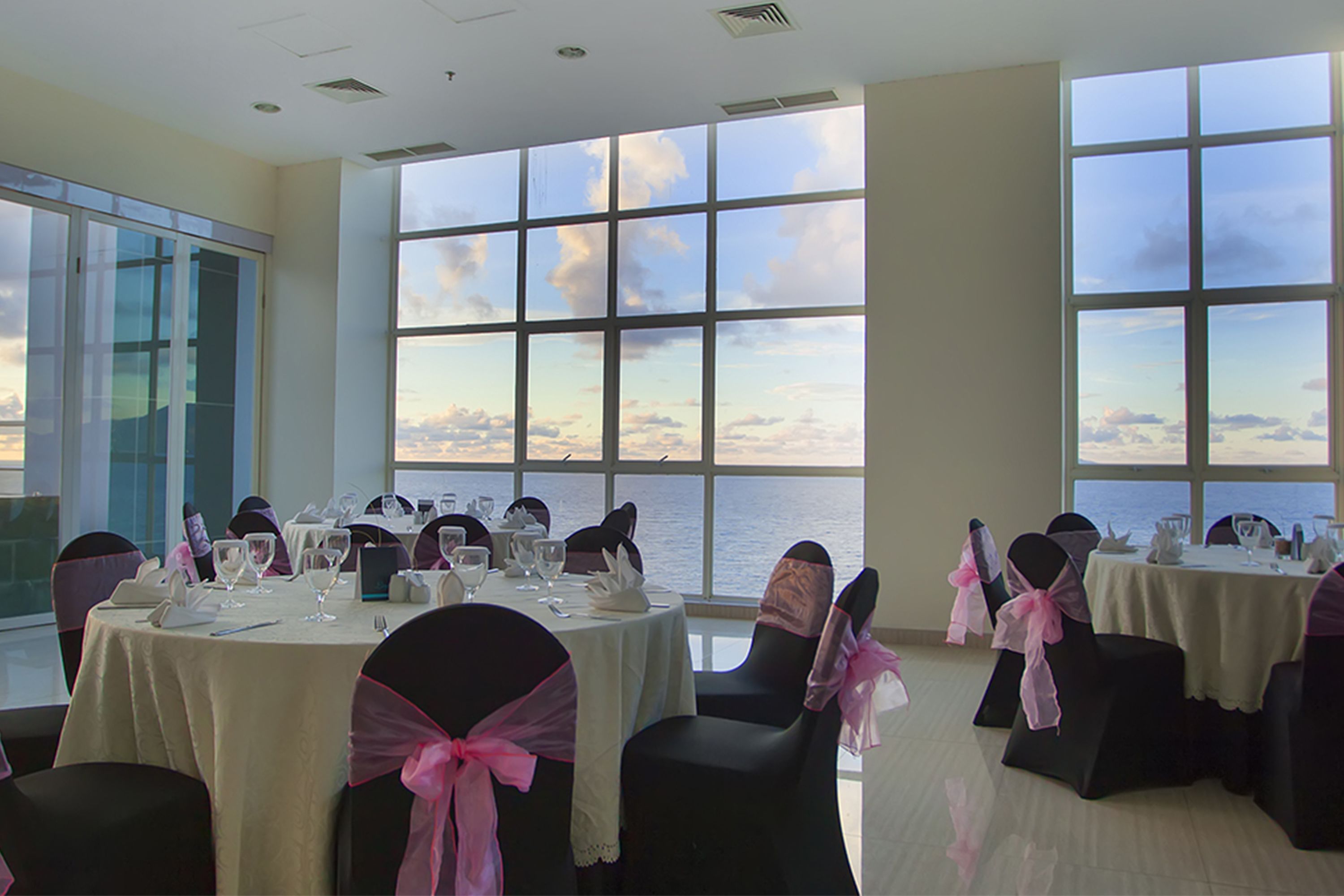 A banquet hall with round tables, chairs with pink sashes, and large windows overlooking the sea.
