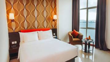 Een hotelkamer met een groot bed, een nachtkastje, een stoel, een tafeltje met een lamp, en uitzicht op het water door het raam.