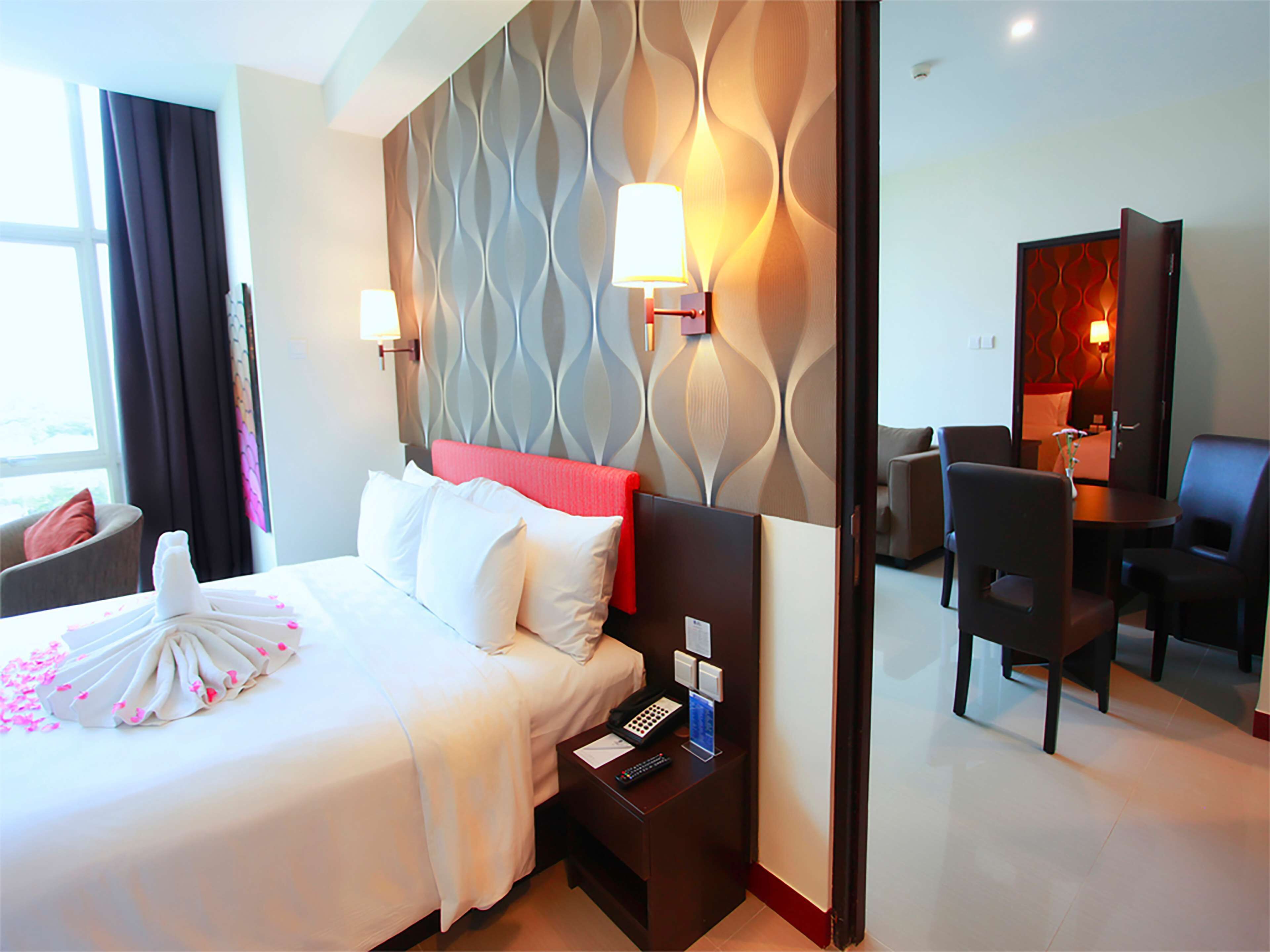 A hotel room with a bed, bedside table, lamp, and a view of the dining area.