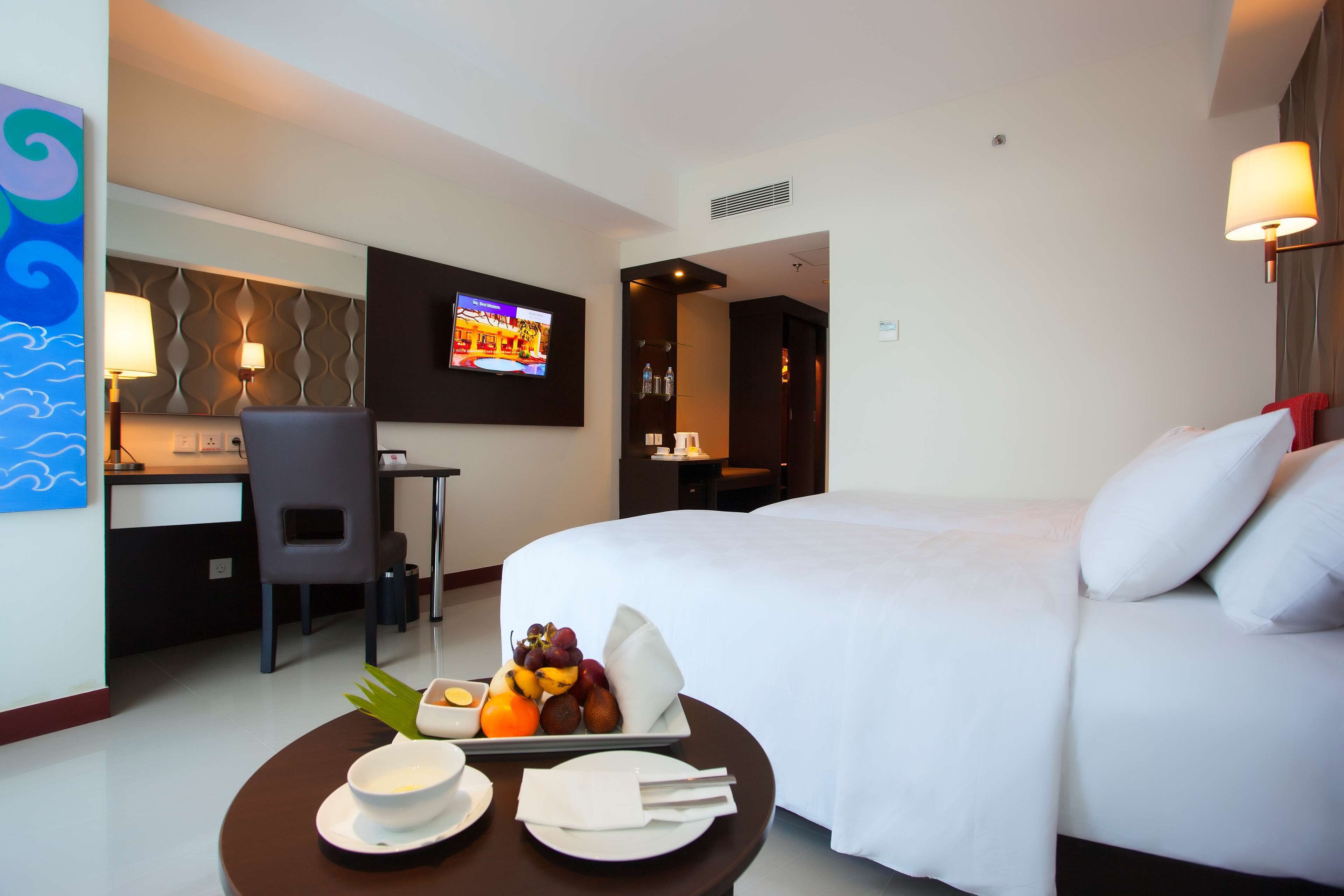 A hotel room with a bed, a desk, a TV, and a breakfast tray with fruits and a bowl.