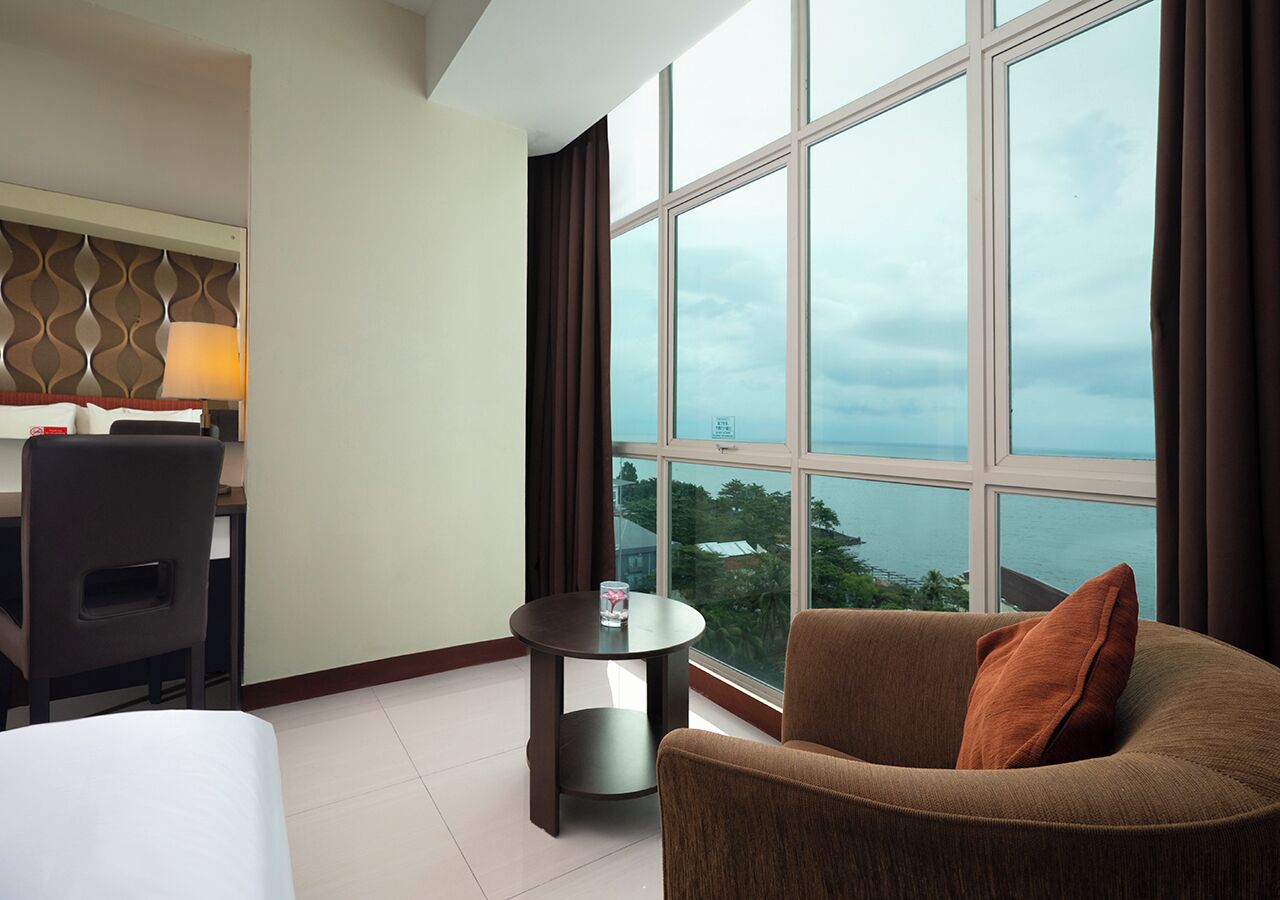 A hotel room with a bed, a desk, a chair, a round table, and a large window with a view of the sea.
