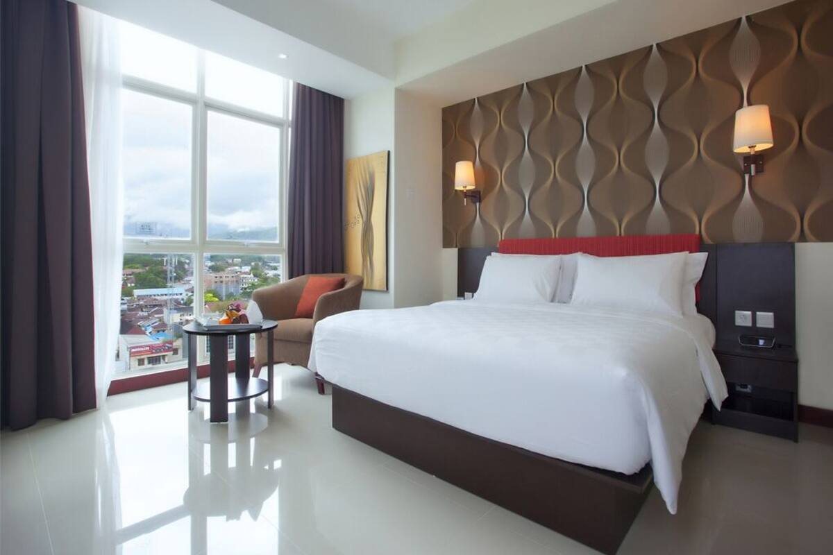 A hotel room with a large bed, a side table, a lamp, a view of the city, and a window with curtains.