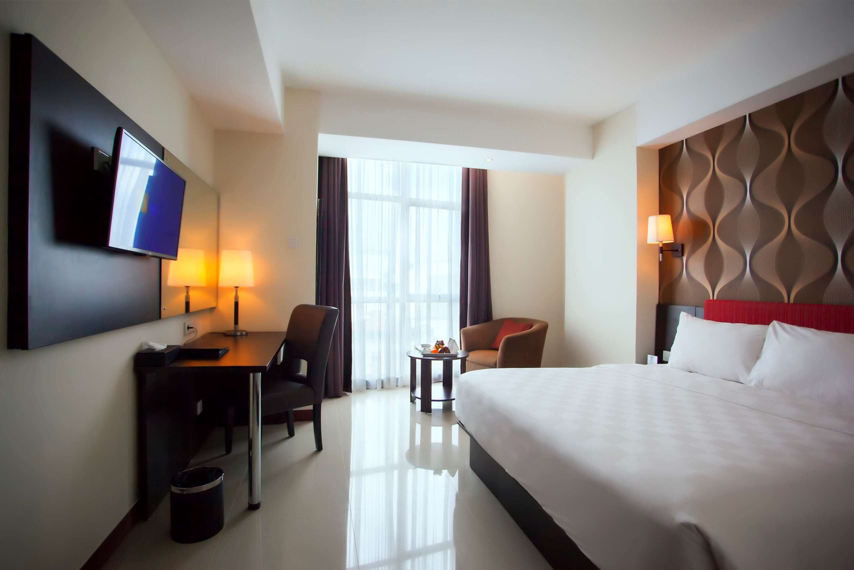 A hotel room with a bed, a desk with a chair, a television, a lamp, and a window with curtains.