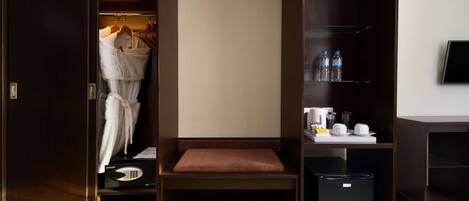 A hotel room with a wardrobe, a seating area, a mini-fridge, and a shelf with toiletries.
