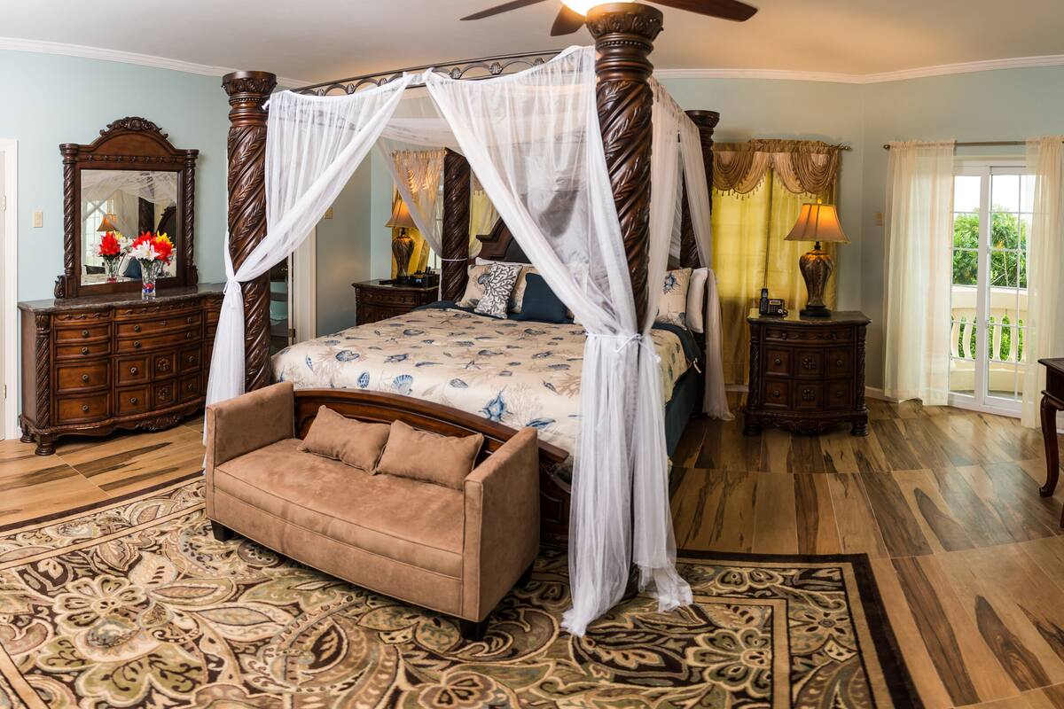 A four-poster canopy bed with a nightstand, a chaise lounge, a desk with a chair, and a large mirror.