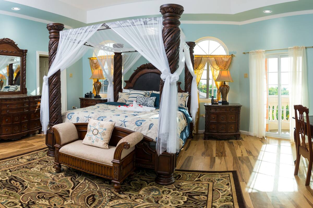A spacious bedroom with a four-poster bed, a sitting area with a chair, a vanity with a mirror, and a dining table with chairs.