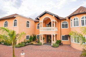 A large, two-story building with a central balcony, surrounded by a courtyard with palm trees and a brick pathway.