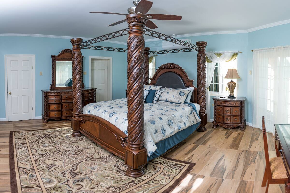 A four-poster bed with a ceiling fan, a nightstand with a lamp, a dresser, and a mirror.