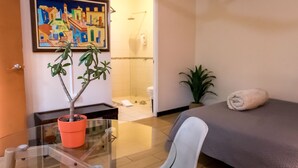 A hotel room with a glass table, a potted plant, a bed with a towel, a painting on the wall, and a shower area. - Terida Airport B&B (Desamparados)