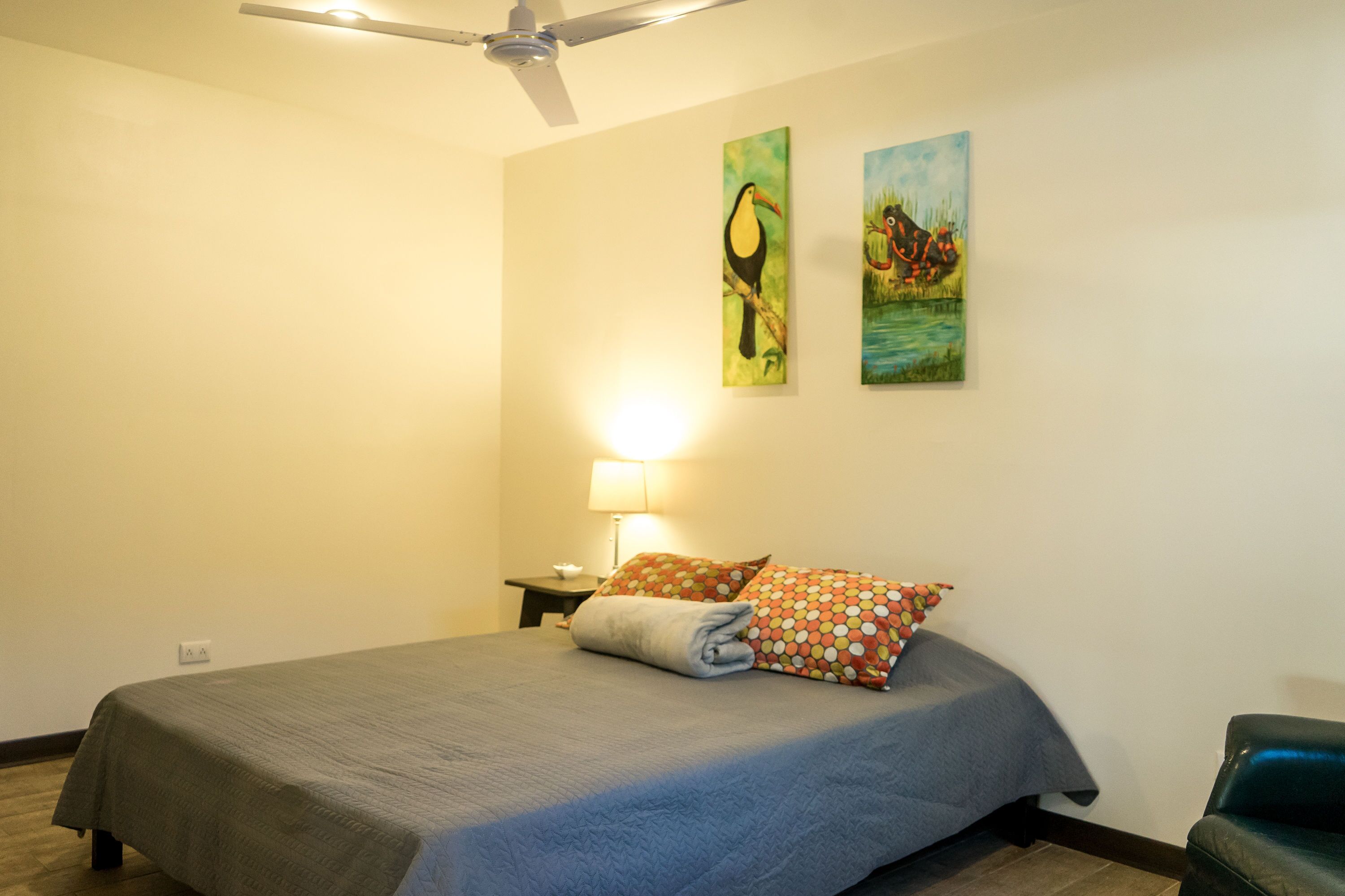 A hotel room with a bed, a ceiling fan, a nightstand with a lamp, and two wall paintings.