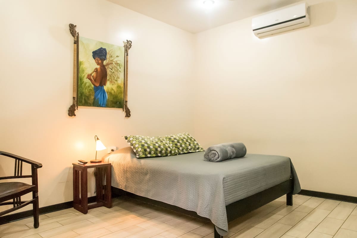 Een slaapkamer met een bed, een stoel, een kleine tafel met een lamp, een schilderij aan de muur en een airconditioner.