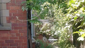 Une ruelle étroite pavée de briques, avec un banc en métal blanc, un pichet en céramique et une plante en pot.