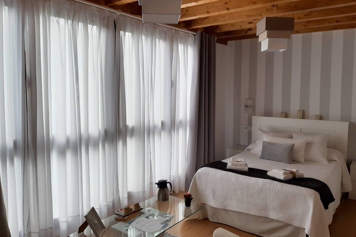 A modern bedroom with a large bed, white curtains, a glass table, and a wooden ceiling.