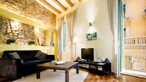 Un salon avec un mur en pierre, un canapé noir, une table basse, un téléviseur et un balcon doté d’une balustrade.
