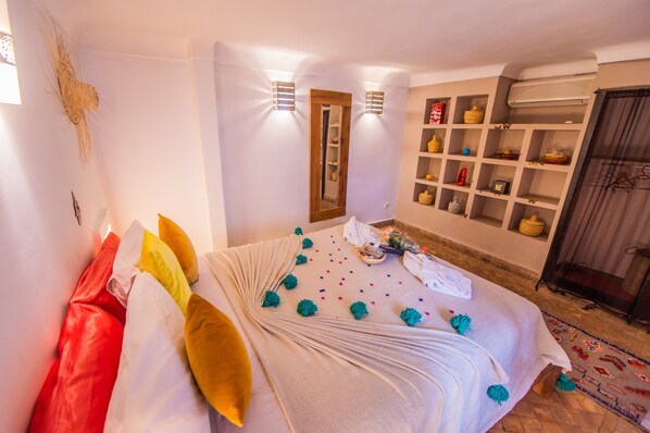 A neatly made bed with colorful pillows and a decorative headboard, a shelf with various items, and a wall-mounted air conditioner.
