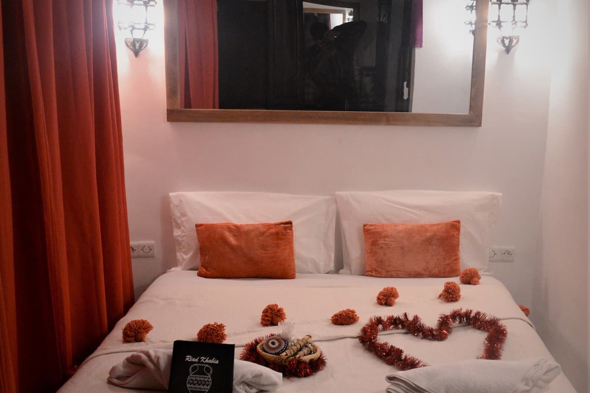 A neatly made bed with orange pillows, a heart-shaped decoration, and a sign on the bed.