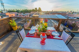 A rooftop terrace with a table set for dining, surrounded by metal railings, potted plants, and a clear view of the cityscape.