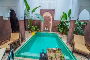 Una piscina con suelo de baldosas, rodeada de plantas en macetas y sillas de madera.
