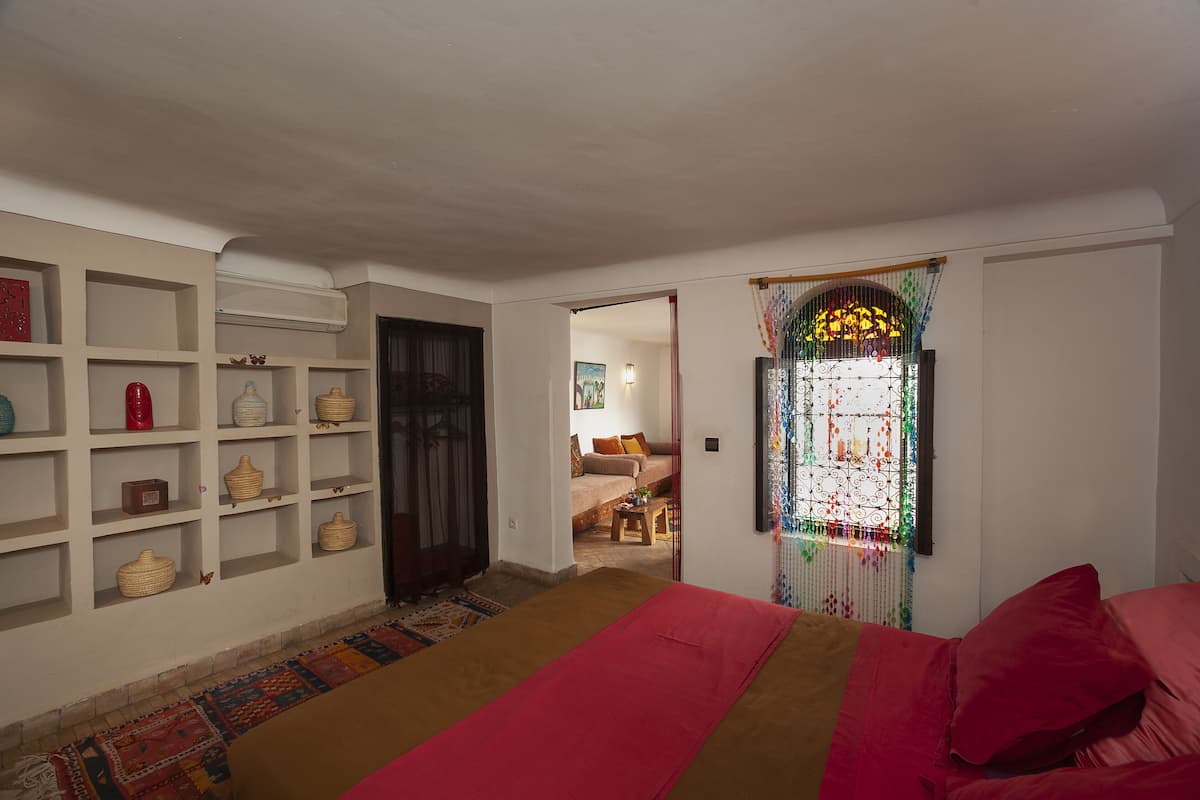 A room with a bed, a shelf with decorative items, a patterned rug, and a doorway leading to another room.